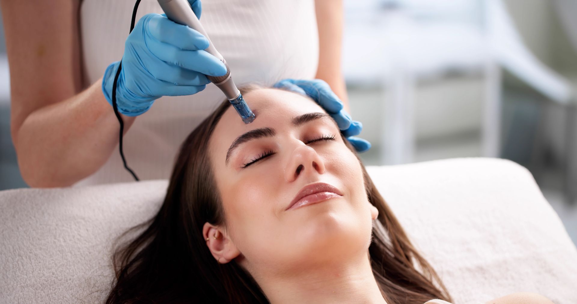 Person receiving facial treatment with tool, indoors. Light skin, blue gloves, eyes closed.