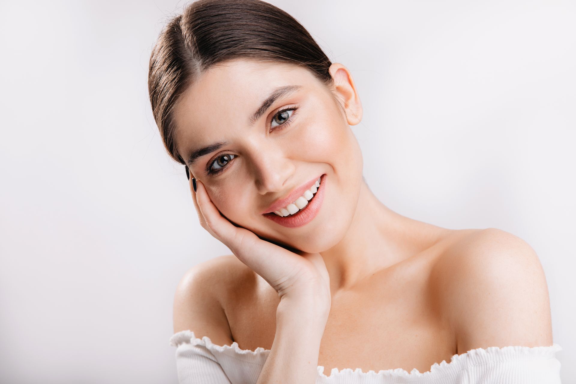Woman with dark hair, smiling, holding her face, wearing an off-the-shoulder white top.