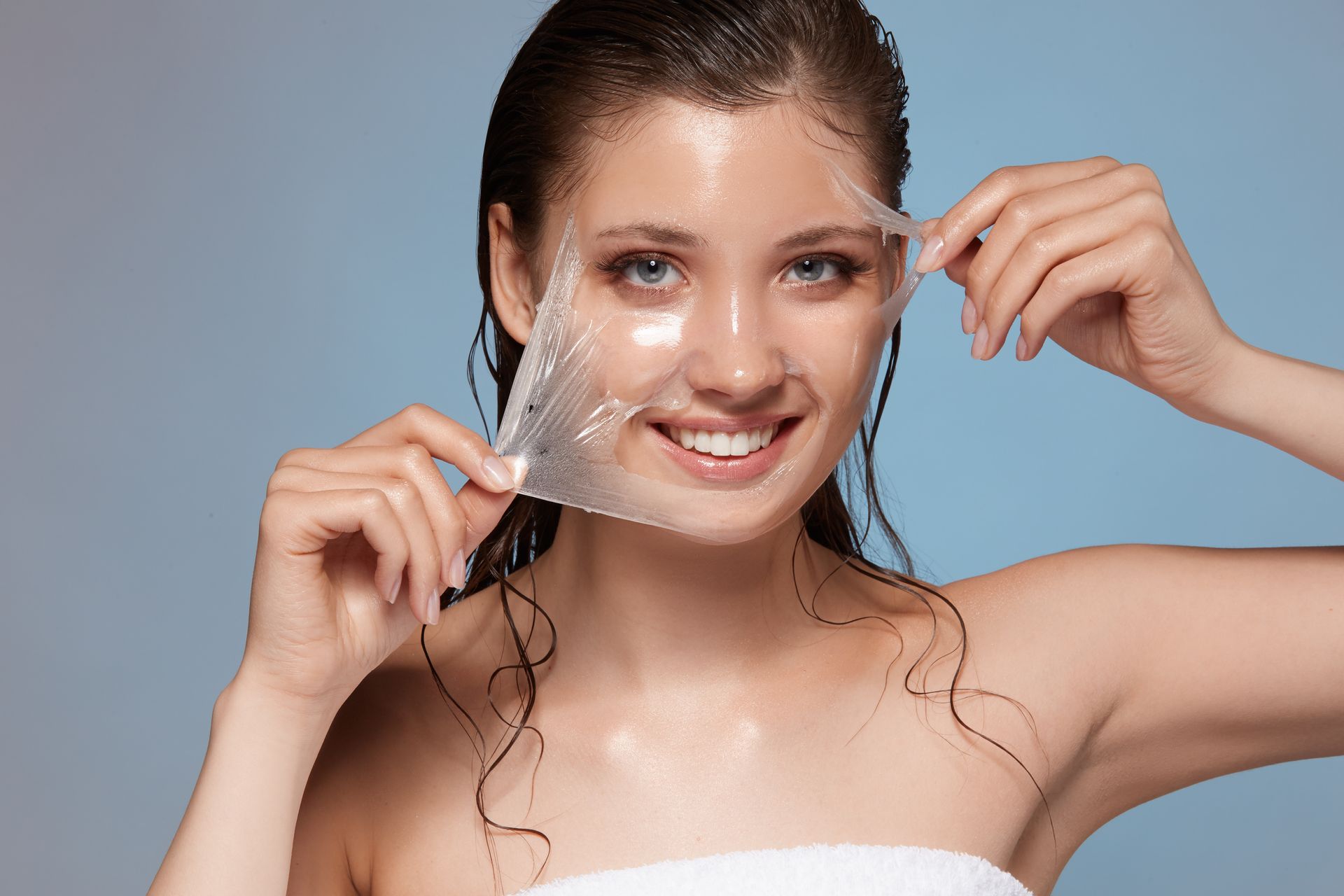 Woman peeling off a facial mask, smiling, against a blue background.