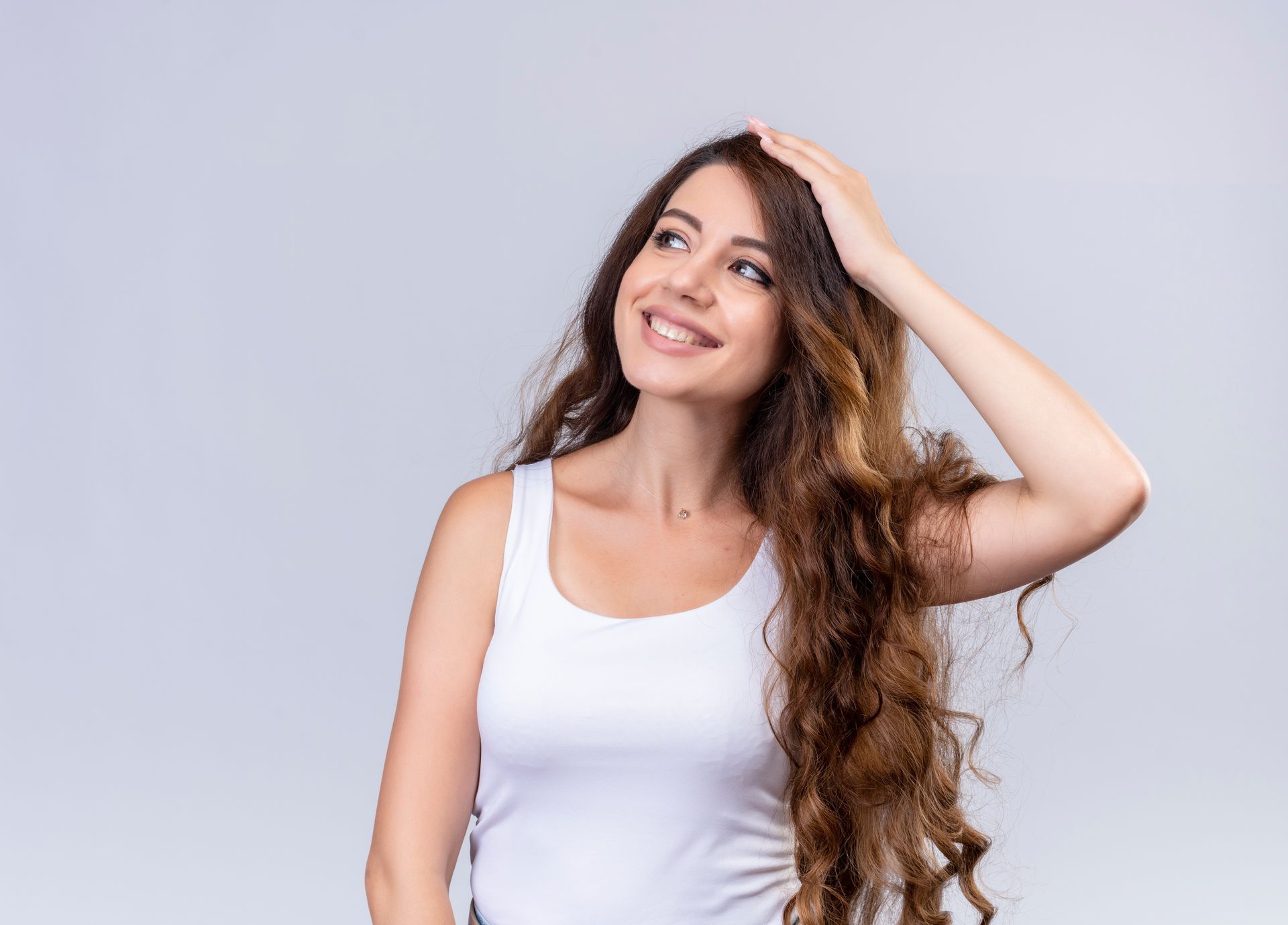 Woman with long, wavy brown hair, wearing a white tank top, smiling and touching her hair, looking up.