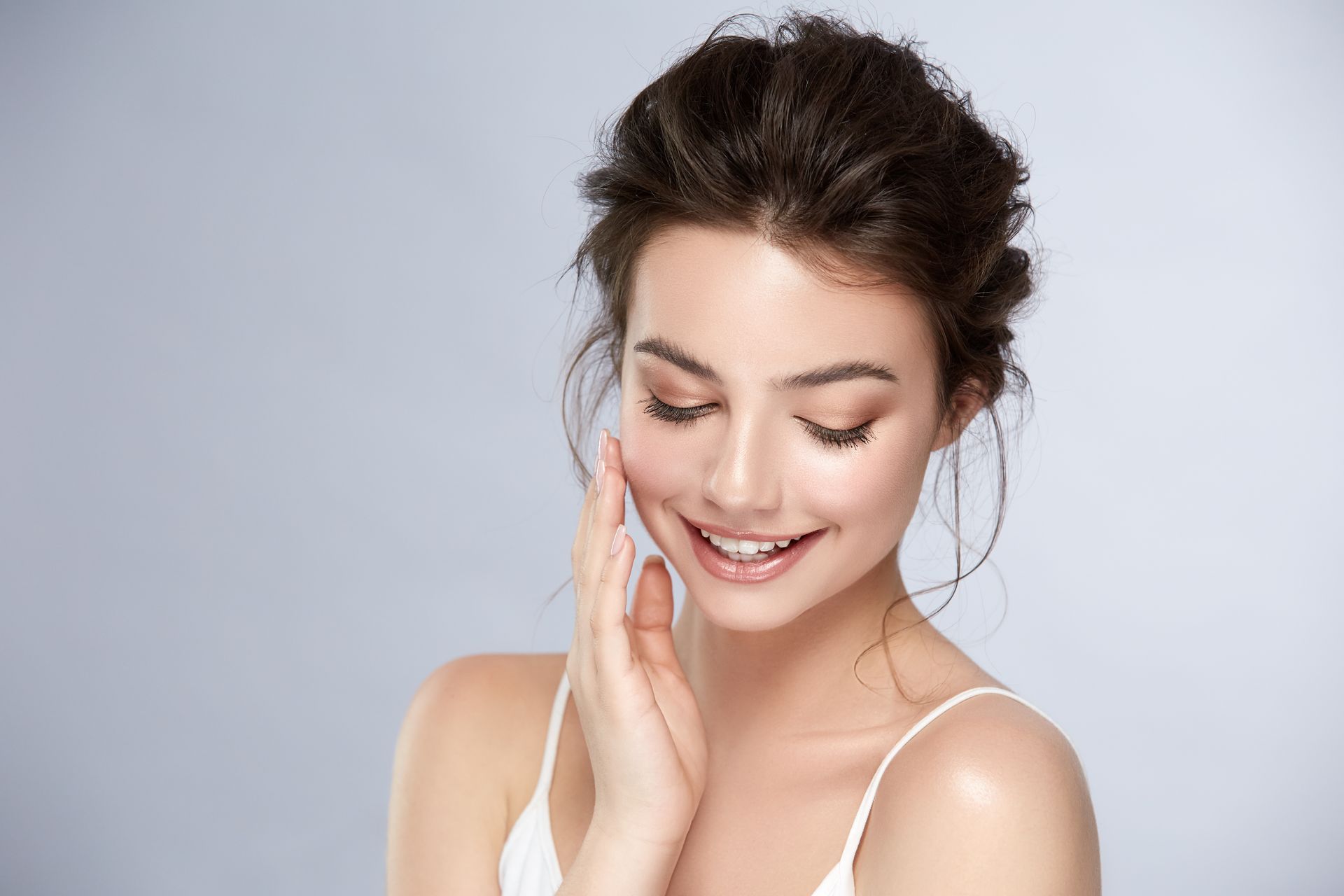 Woman smiles, touches her cheek, wearing a white tank top. Messy updo, neutral background.