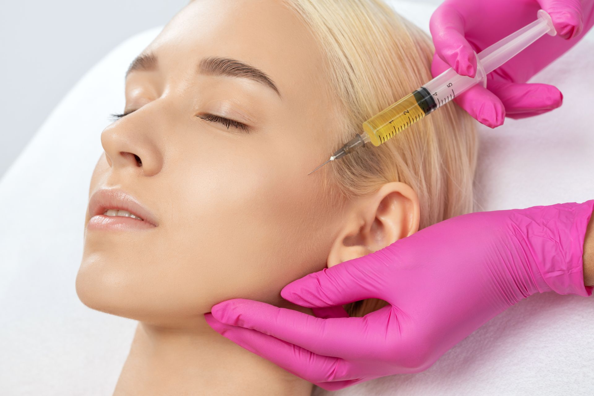 Gloved hands inject facial serum into a reclining woman’s forehead at a spa treatment