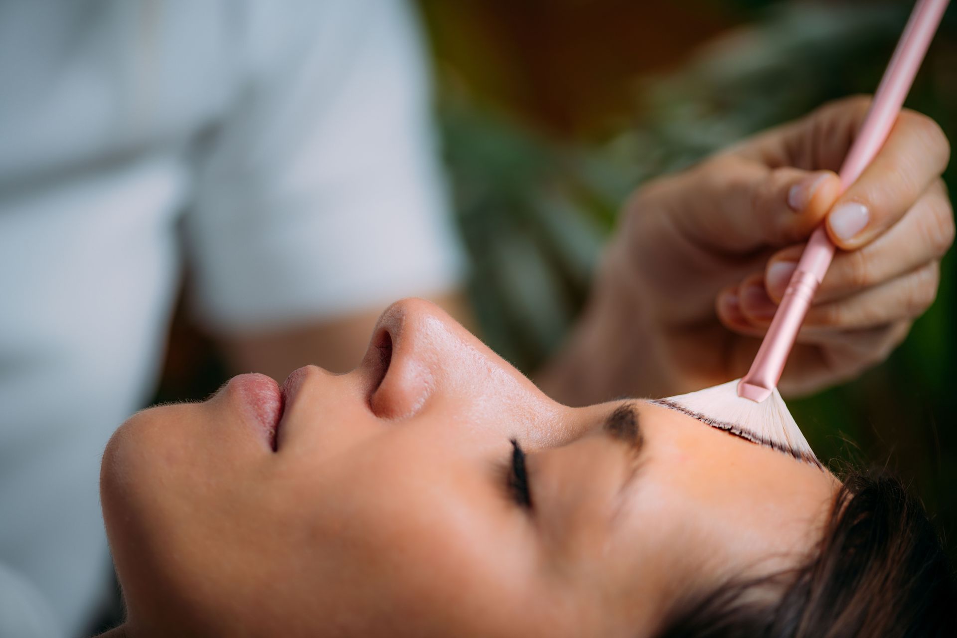 Cosmetician applying a pink facial mask with a brush to a client lying down