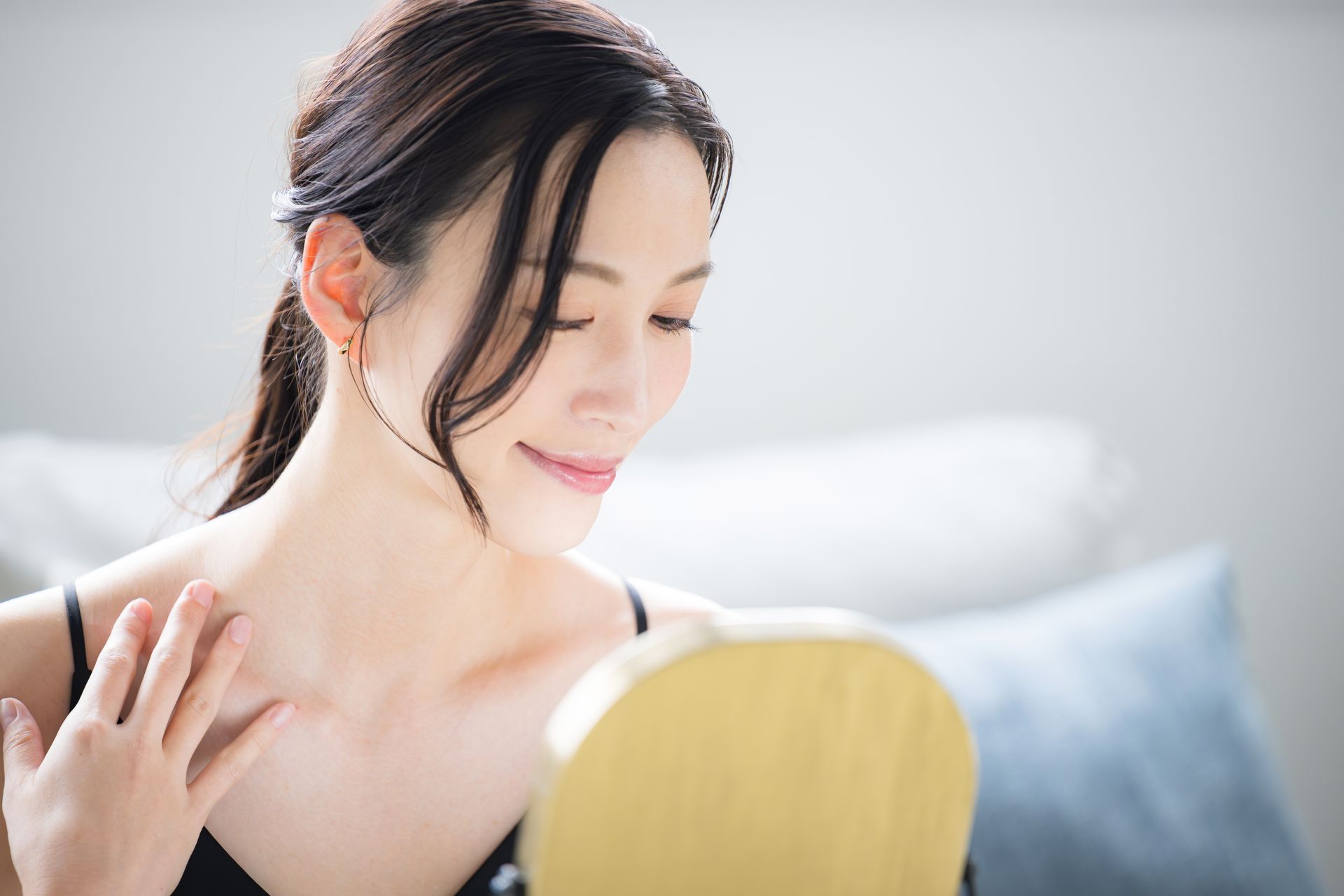 Woman looking in a mirror, touching her neck. She wears a black top, has dark hair, and is smiling in a bright setting.