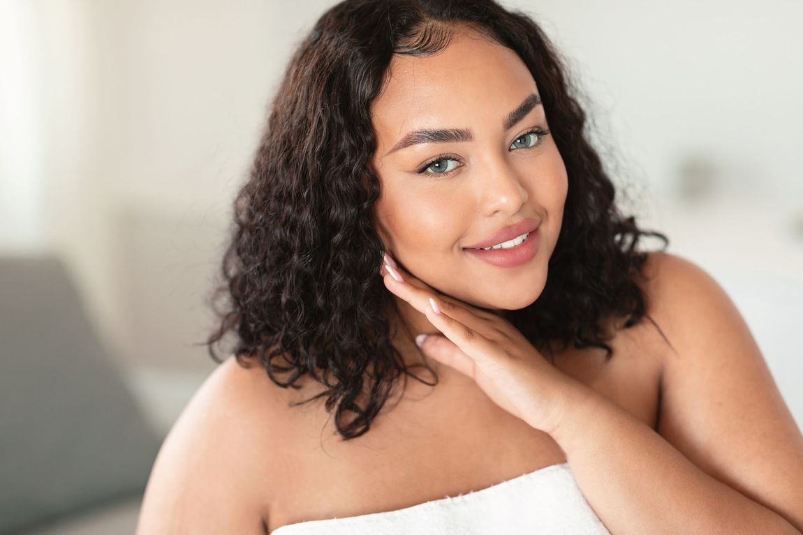 Woman with curly hair touches her neck, smiling. Wearing a white towel.