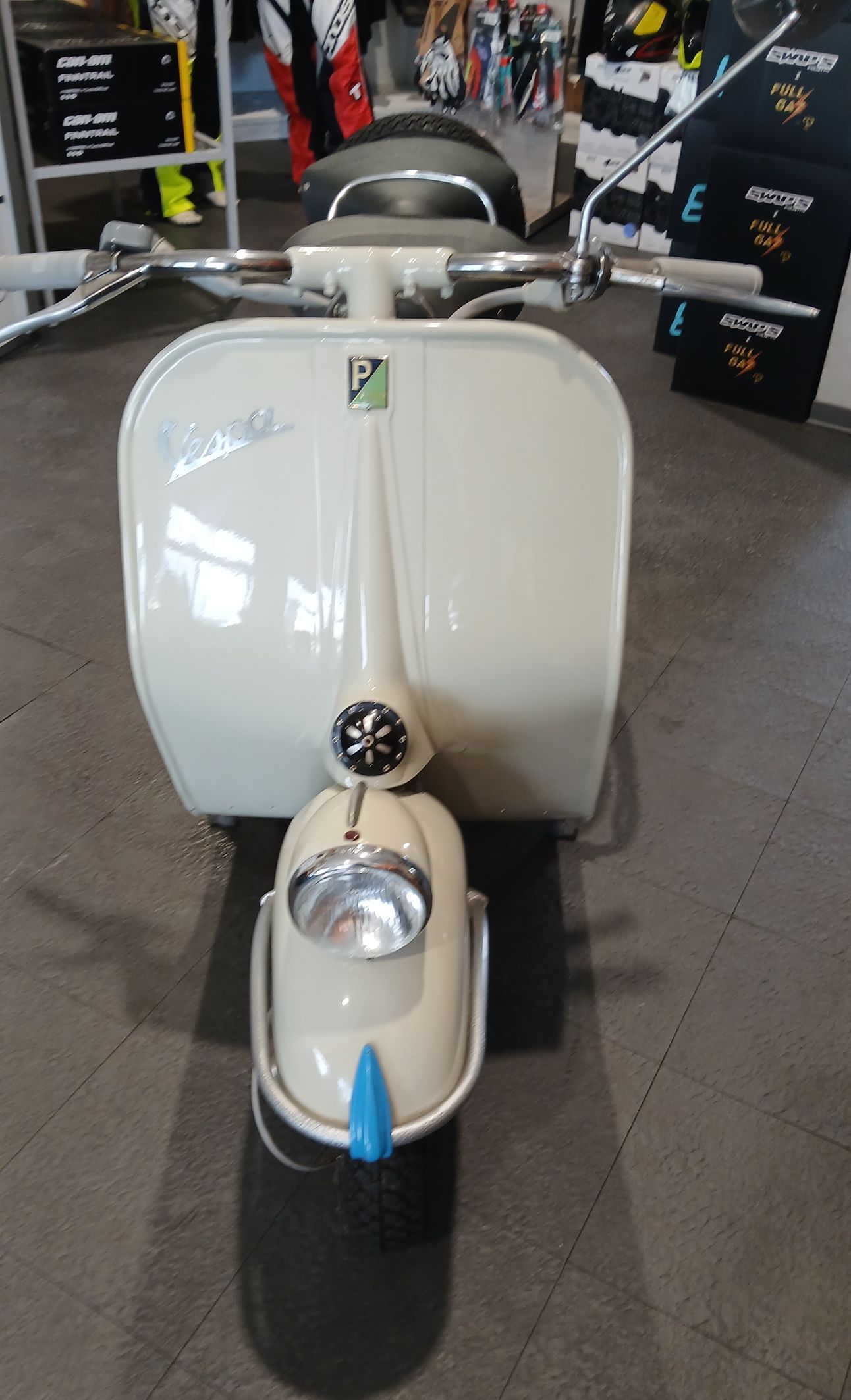 Scooter Vespa color crema visto frontalmente, in un ambiente interno. Manubrio e faro anteriore argentati, finiture blu.