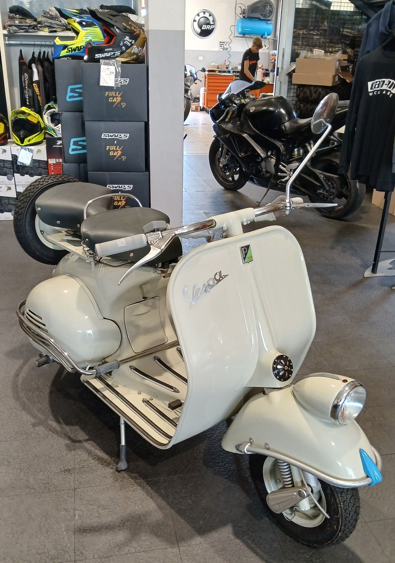 Uno scooter Vespa d'epoca color crema parcheggiato al chiuso, con una moderna motocicletta nera sullo sfondo.