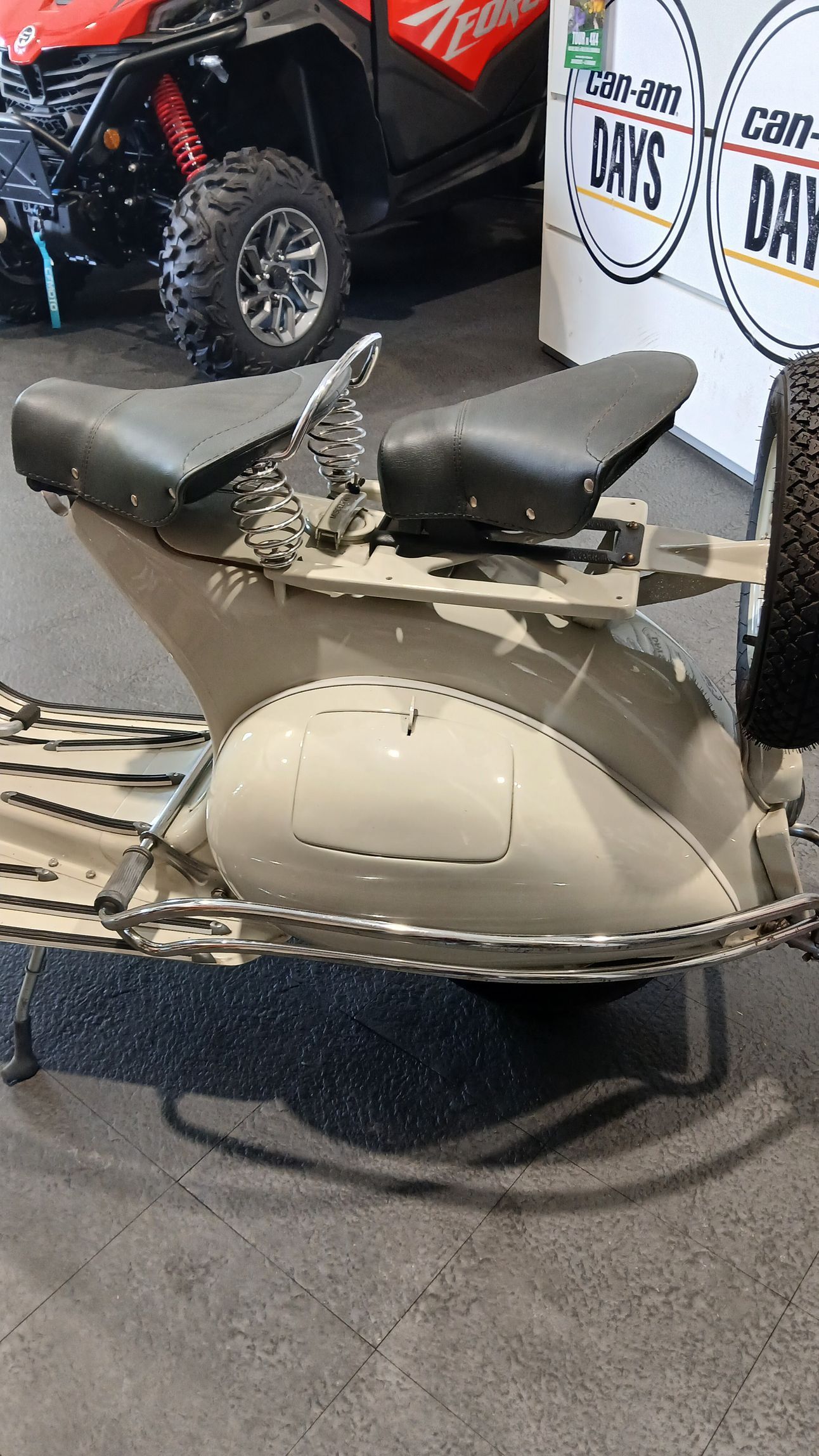 Uno scooter d'epoca con due sedili neri e carrozzeria grigio chiaro. In uno showroom accanto a un fuoristrada.