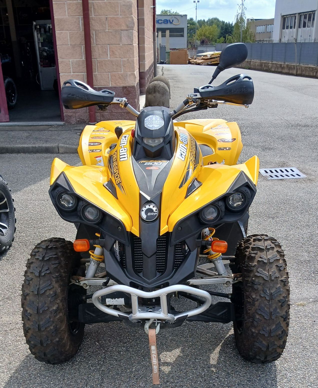 ATV Can-Am giallo con dettagli neri parcheggiato sull'asfalto. Pneumatici fangosi, paramani e cinghia di traino visibili.