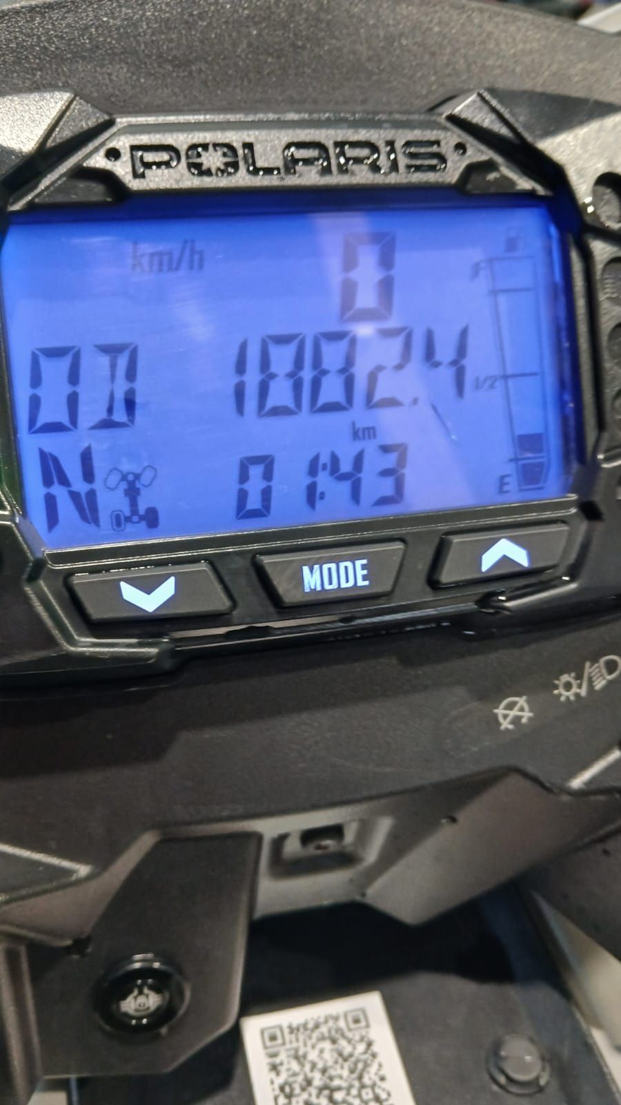 Display digitale Polaris che indica 0 km/h, chilometraggio 1002,4 km e 01:43. Pulsanti neri e sfondo neutro.
