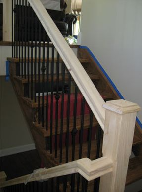 Staircase with wooden steps, black metal spindles, and unfinished wooden handrails, blue tape on edges.