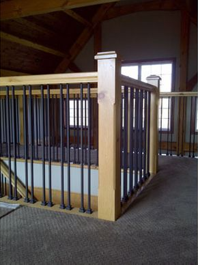Loft railing with black vertical rods and light wood posts, carpeted floor, windows in background.