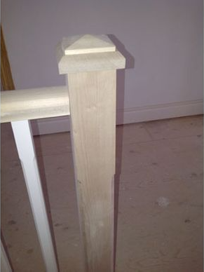 Wooden stair post with a pyramid-shaped top, next to a white railing on a light wood floor.