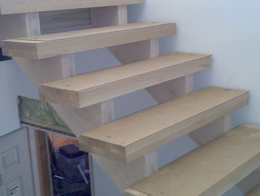Wooden staircase with open risers, ascending against a white wall.