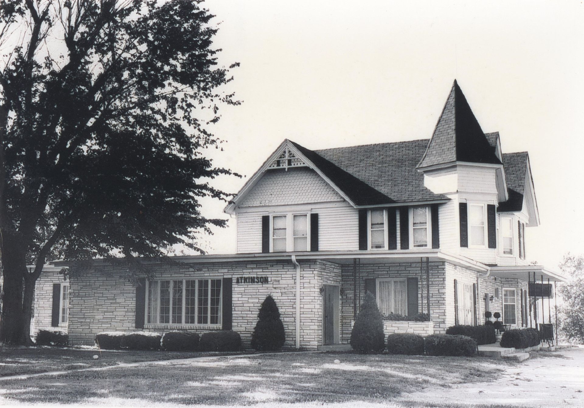 Black and white photo of a two-story house with a small tower and wrap-around porch.