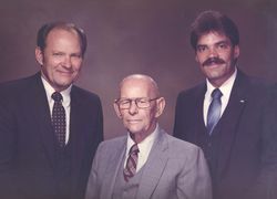 Three men in suits posing for a formal portrait. The older man in the middle, others flanking him.