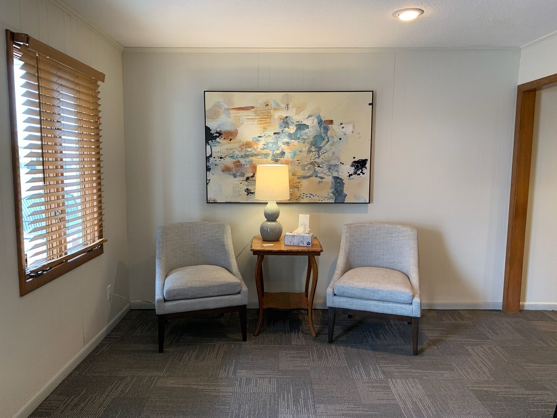 Two chairs face each other in a room with artwork and a side table with a lamp, tissue box, and window with blinds.