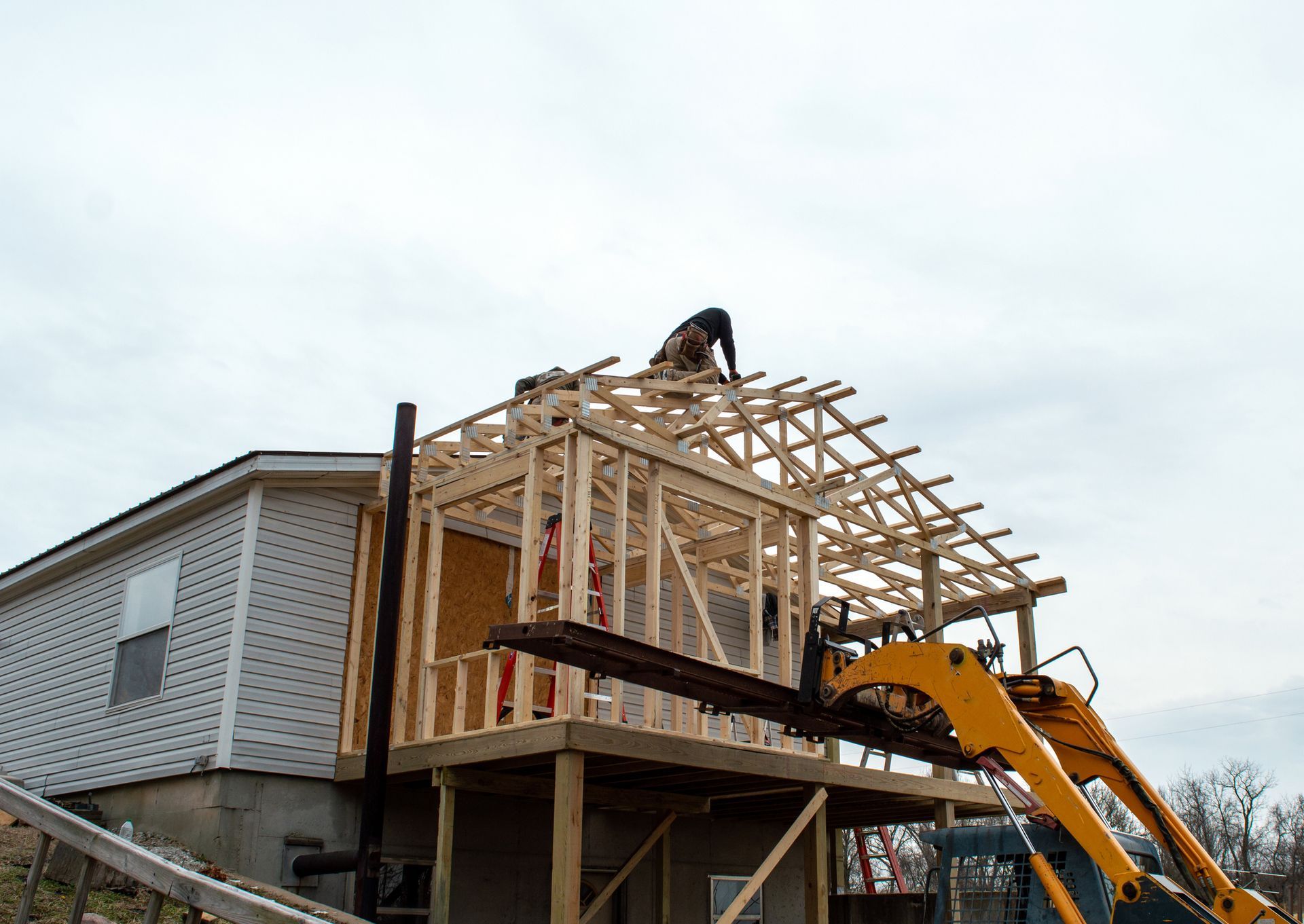 Construction worker building a wooden roof frame on a house extension with a yellow excavator.