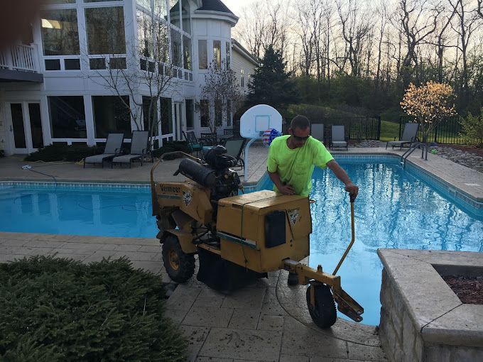 A professional tree trimmer operating a stump grinding machine next to a swimming pool beside a house.