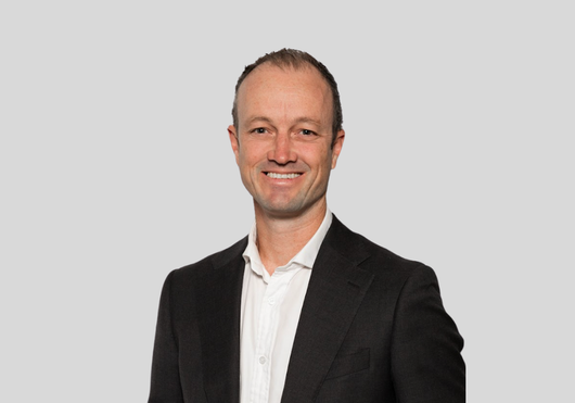 A person with a neutral expression wearing a dark suit jacket and a white button-down shirt against a light gray background.