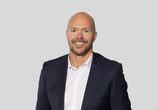 A smiling person with a shaved head and a beard, wearing a white shirt and navy blue suit jacket against a gray background.