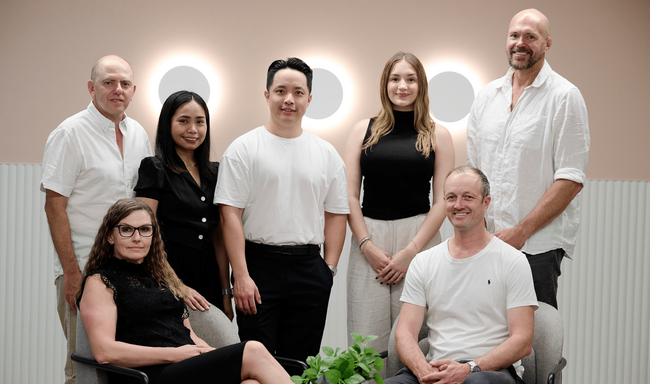 Seven people posed together in a light-filled office space with a neutral background and wall-mounted circular lights.