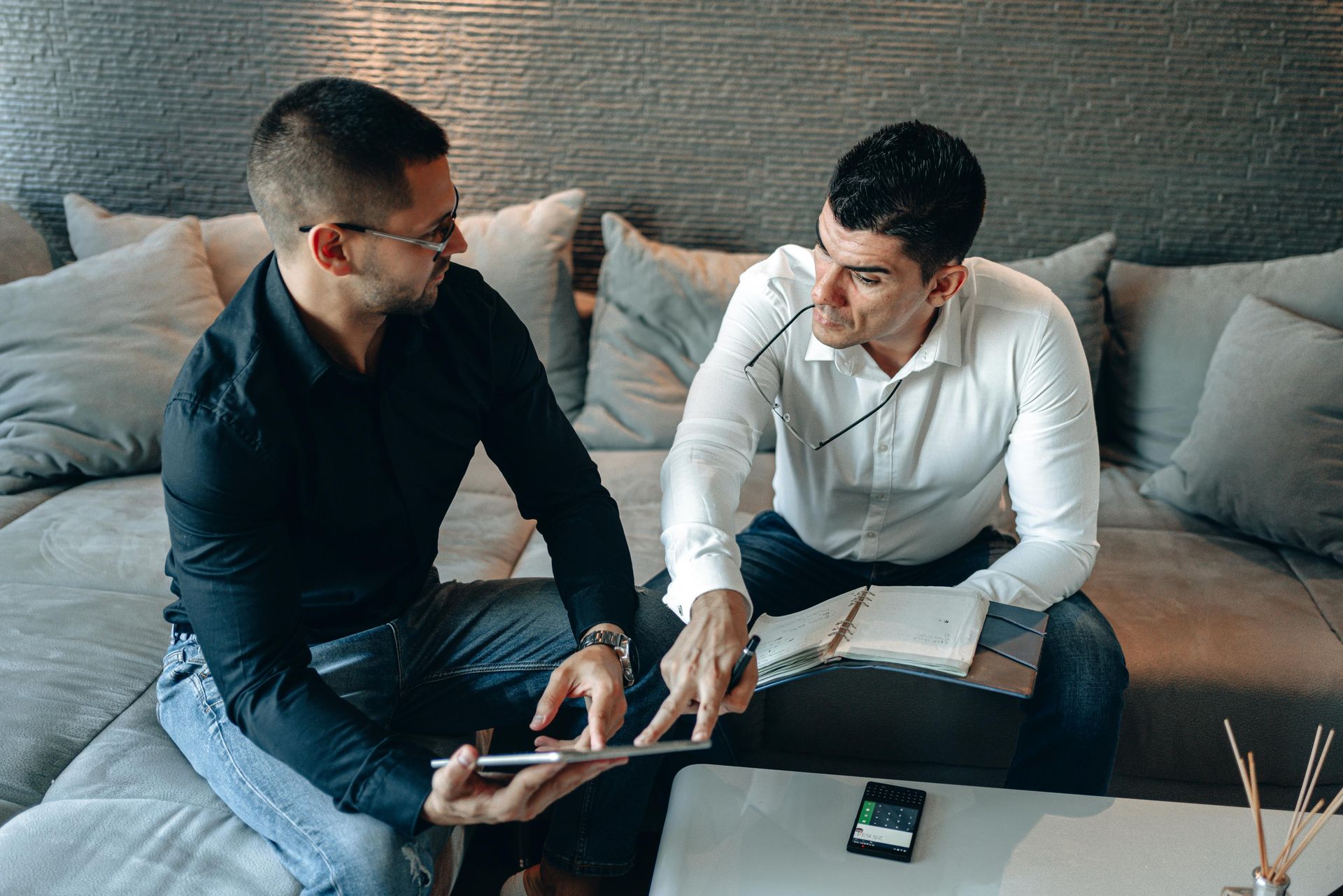 Two men discussing documents on a couch with a laptop and phone nearby