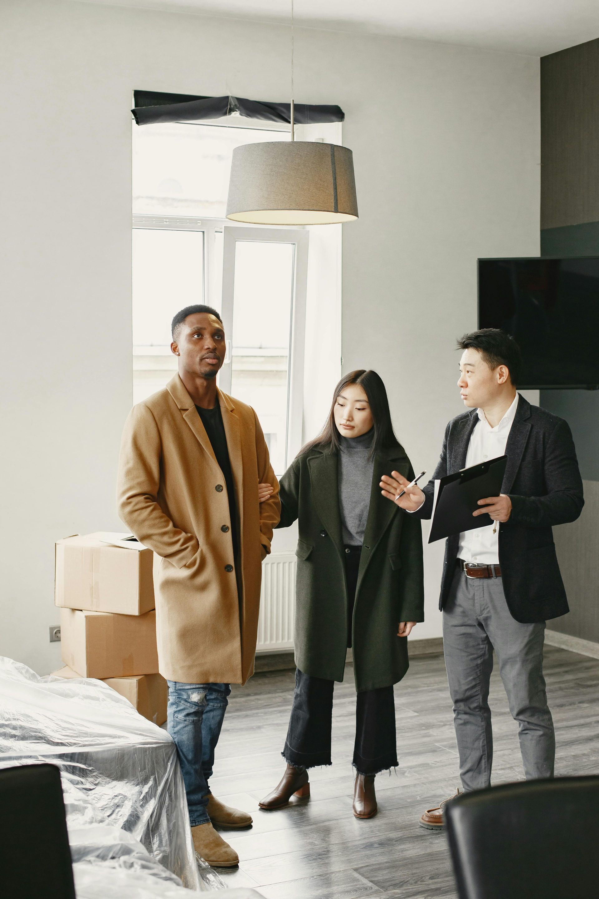 Three people standing in a bright living room, talking near windows and moving boxes.