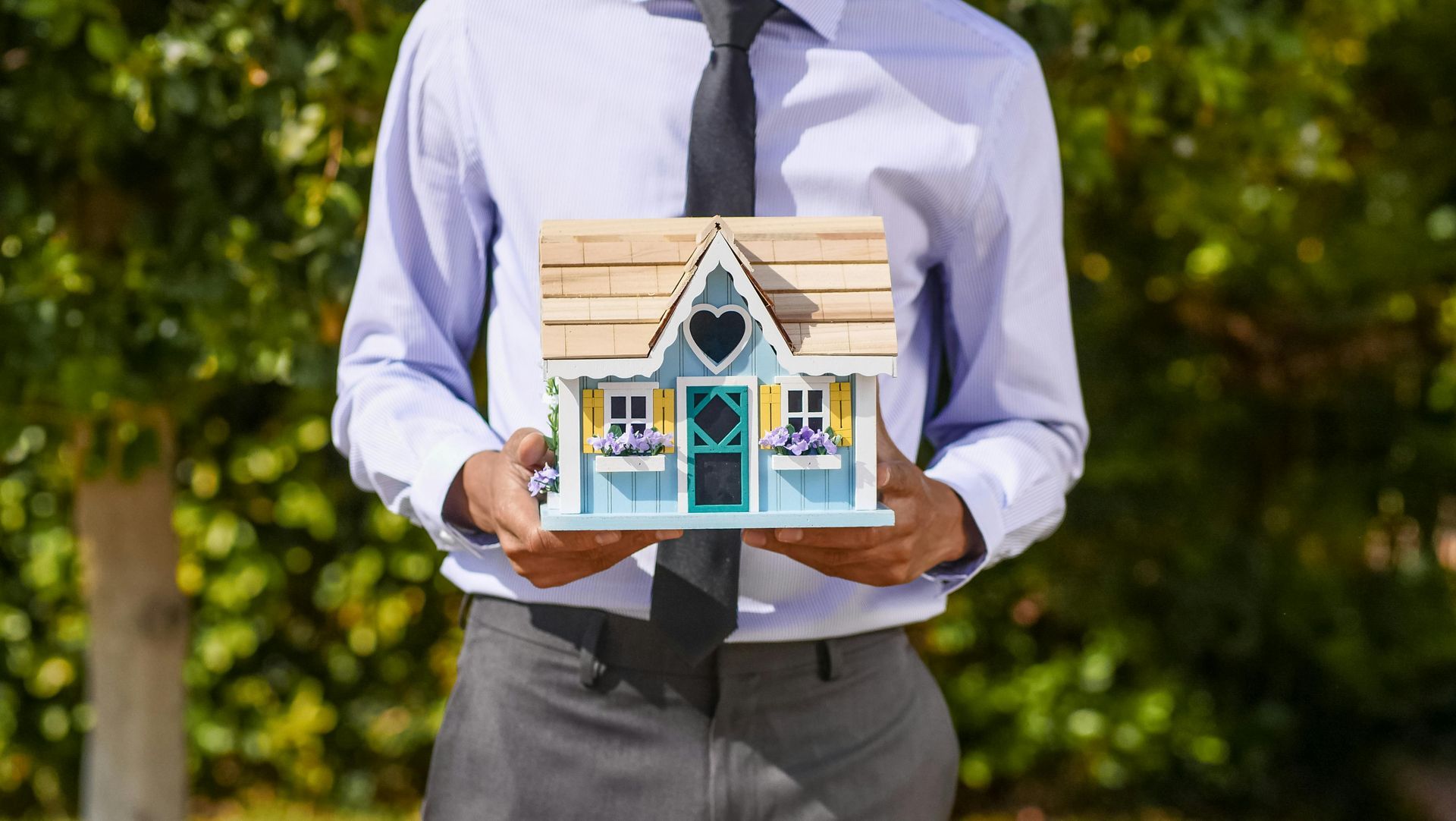 Person holding a small blue house model outdoors in a shirt and tie