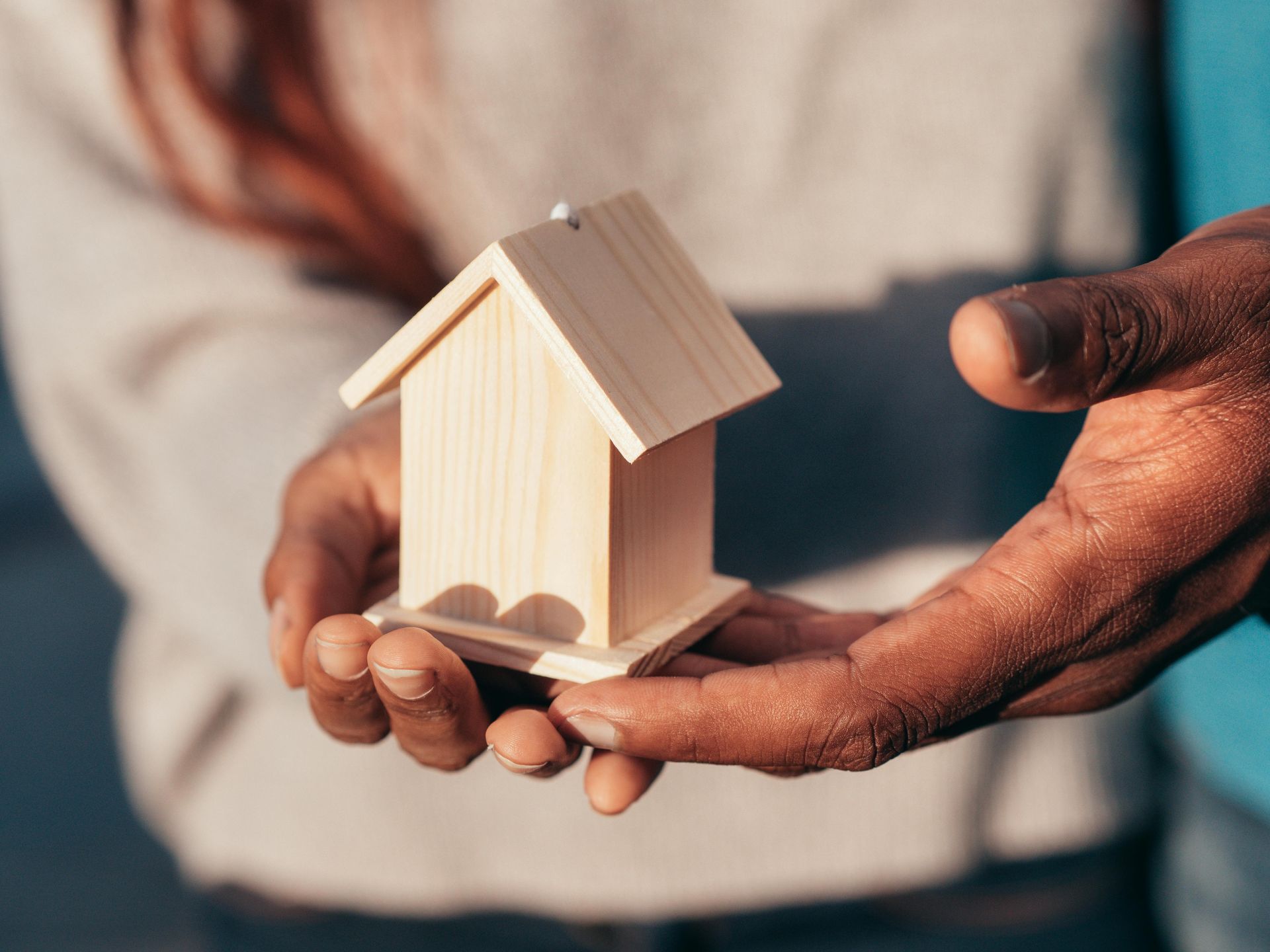 Hands gently holding a small wooden house model with a teal background