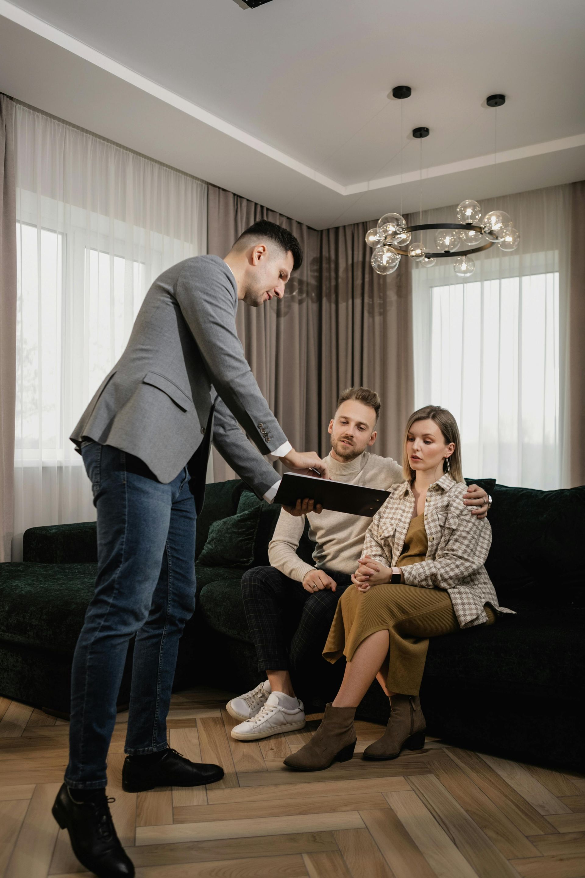 Three people in a living room, one handing a card while two sit on a black sofa