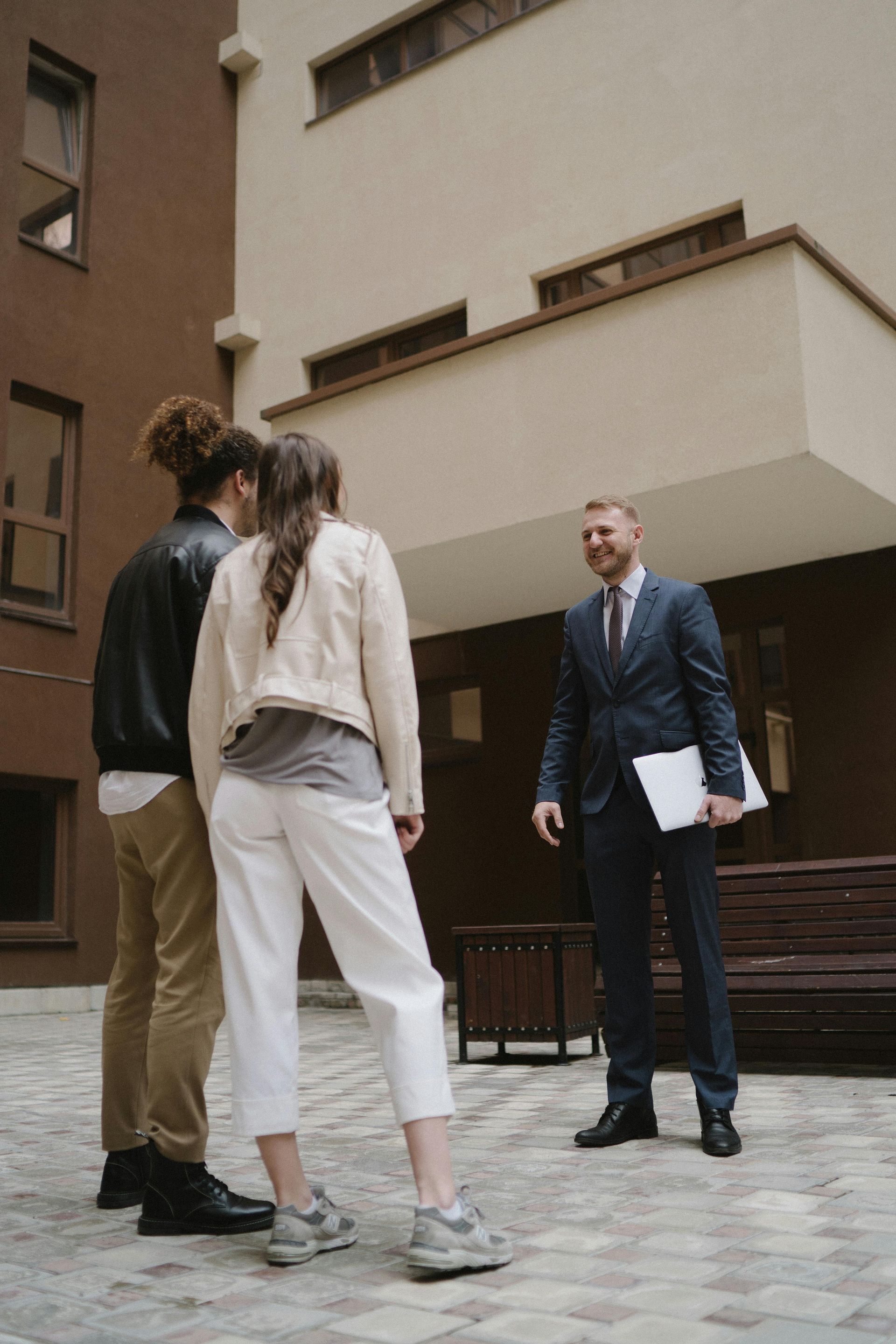 Three people standing in an apartment courtyard, speaking with a man in a suit holding papers.