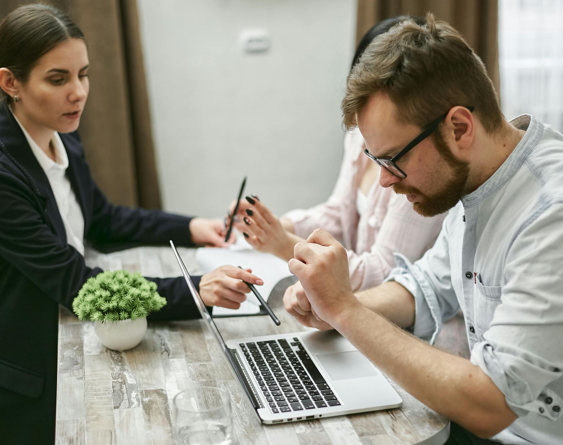 Three coworkers discuss documents around a laptop at a table in an office