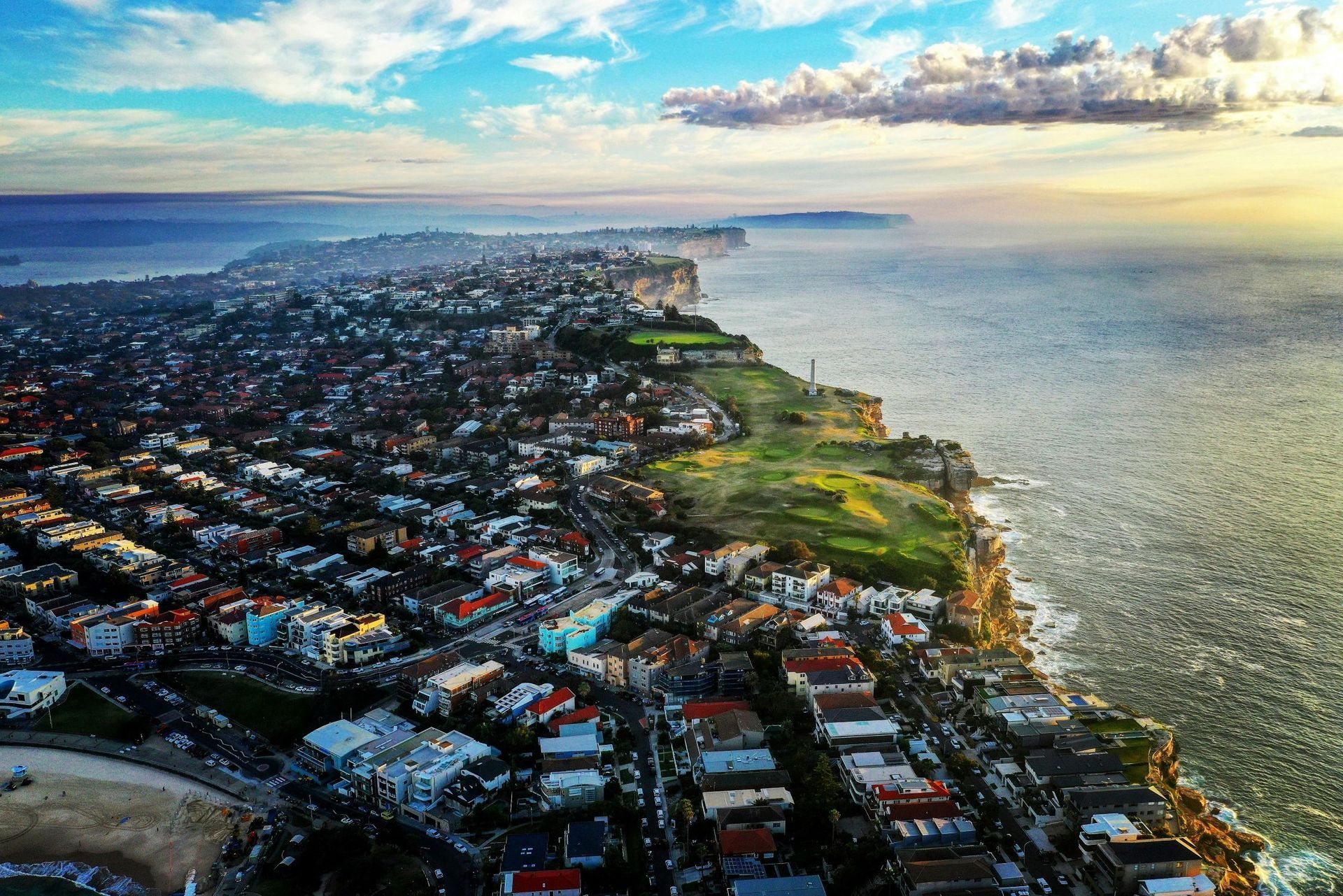 Coastal town on a cliffside beside the ocean at sunset, with dense buildings and blue sky.
