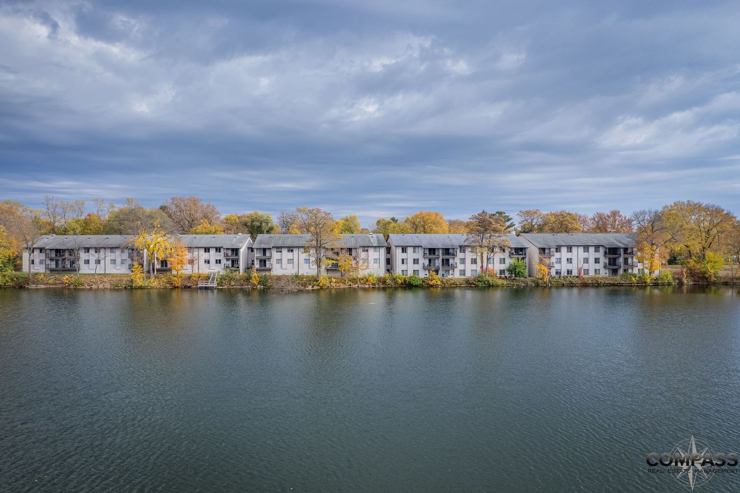 Row of lakeside apartments with autumn trees beneath a cloudy sky, reflected in calm water
