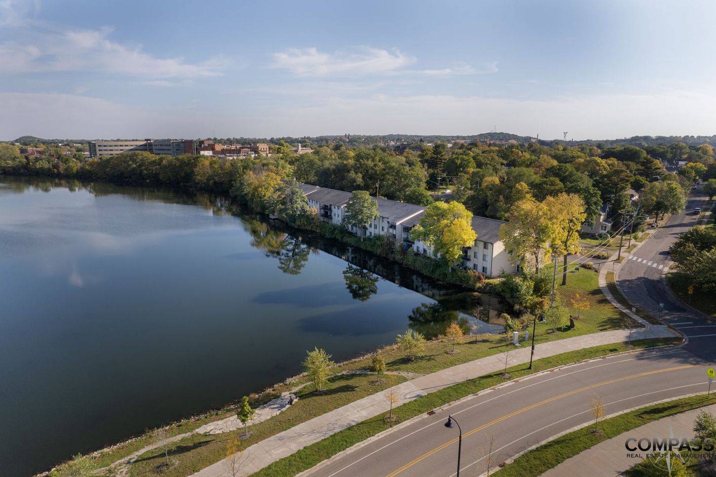 Aerial view of a lakeside road, trees, and houses on a sunny day.