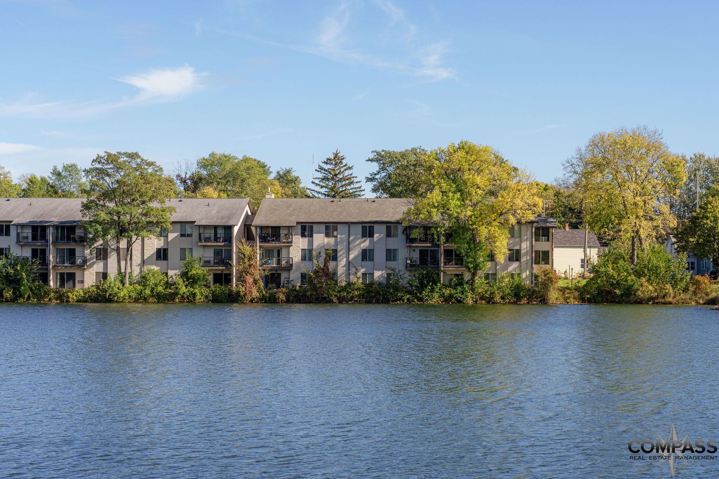 Lakeside apartment buildings with trees reflected in calm water under a clear blue sky