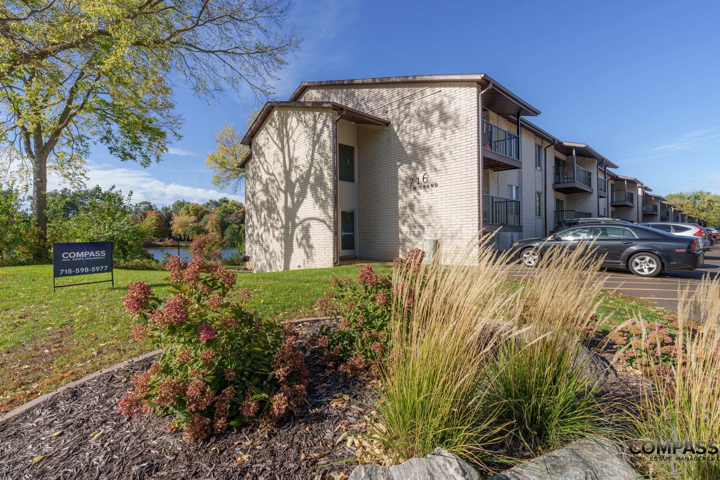 Apartment building entrance with landscaped shrubs, grass, and parked cars on a sunny day