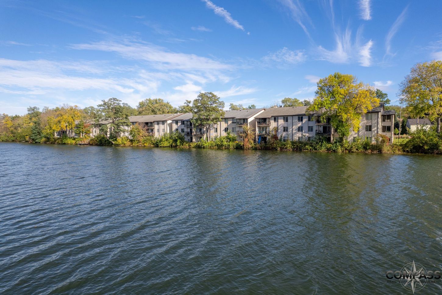 Lakeside apartment buildings behind calm water under a blue sky with wispy clouds