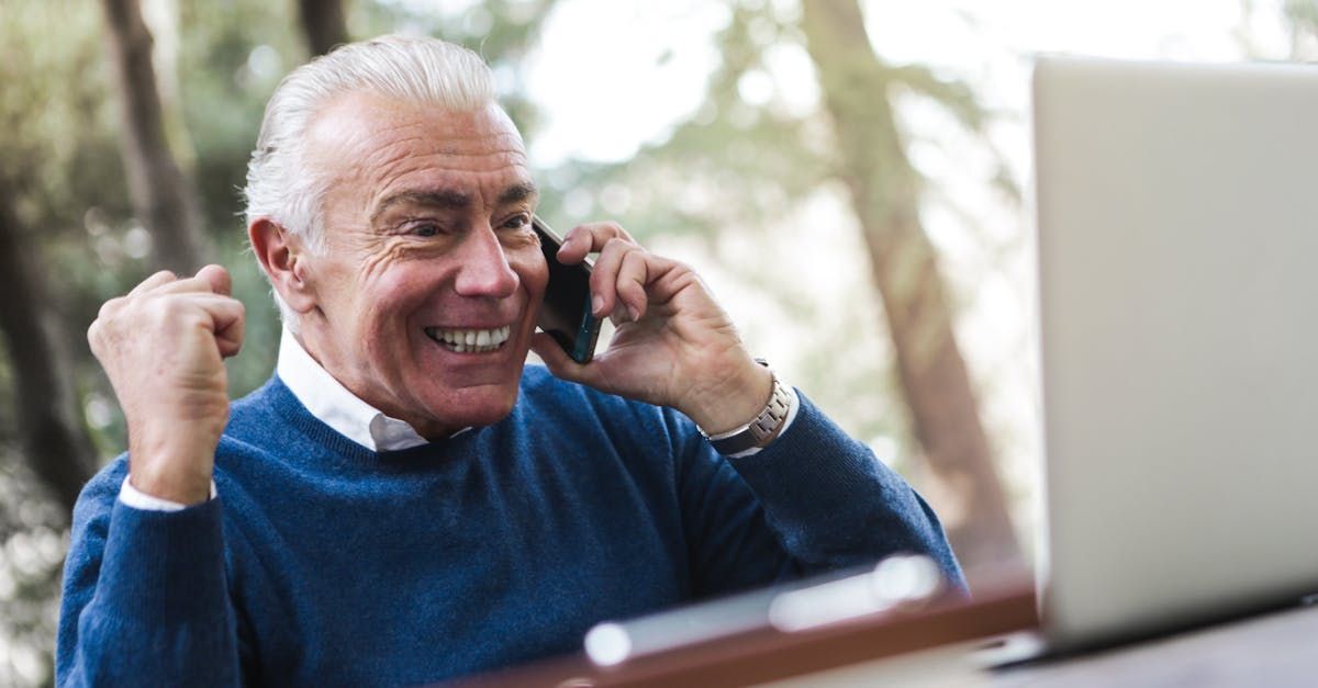 An elderly man is sitting in front of a laptop computer talking on a cell phone.