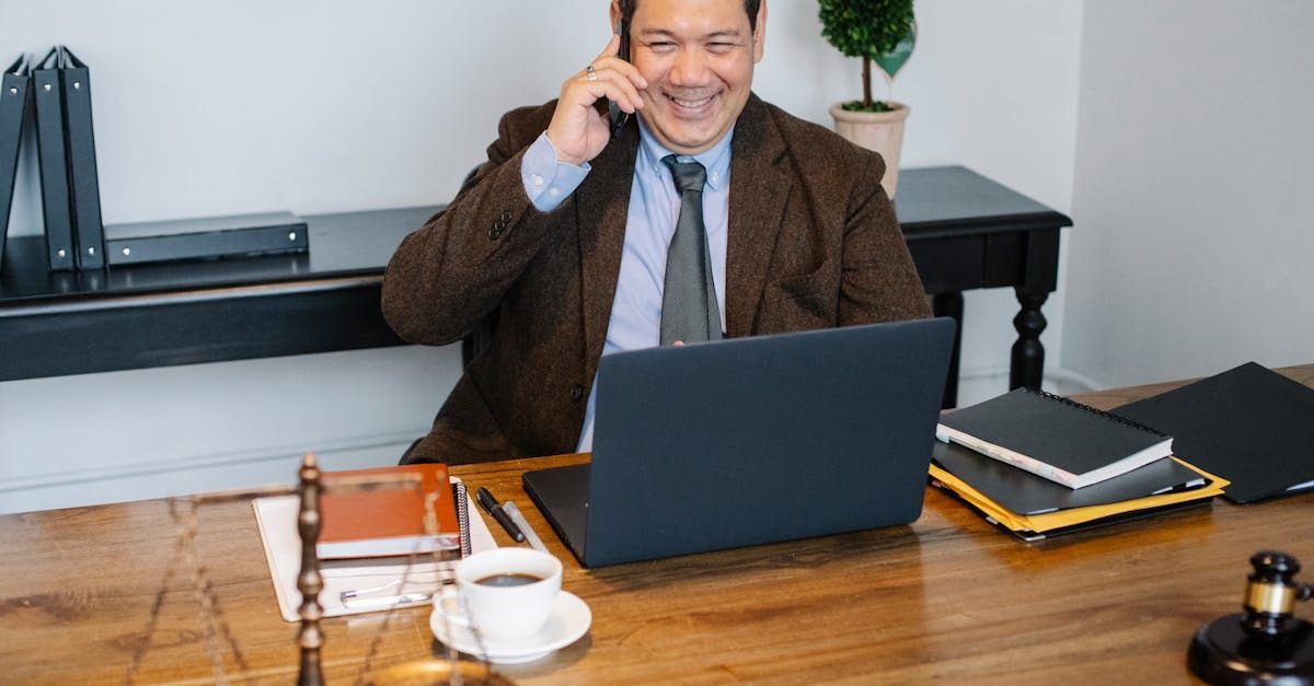 A man is sitting at a desk talking on a cell phone in front of a laptop computer.