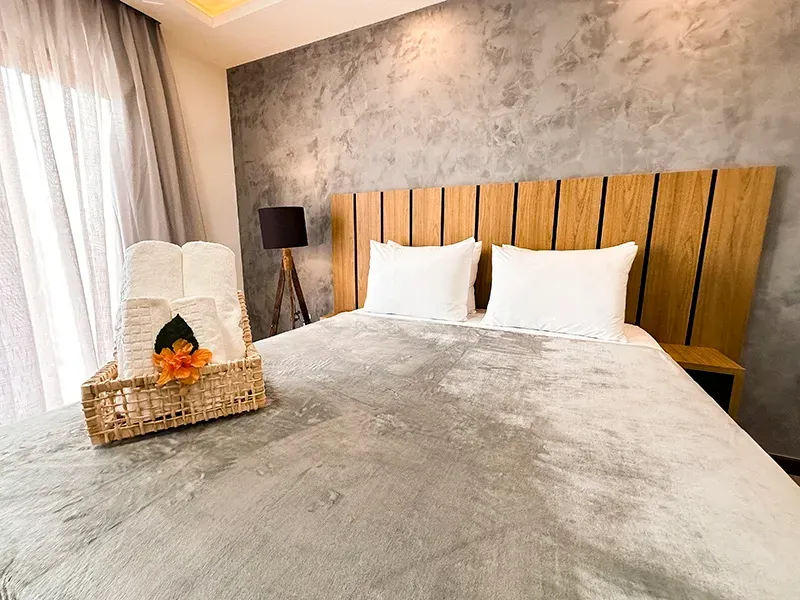 Uma cama de hotel com parede texturizada em tons de cinza, cabeceira de madeira, dois travesseiros brancos e uma cesta de toalhas com uma flor laranja.