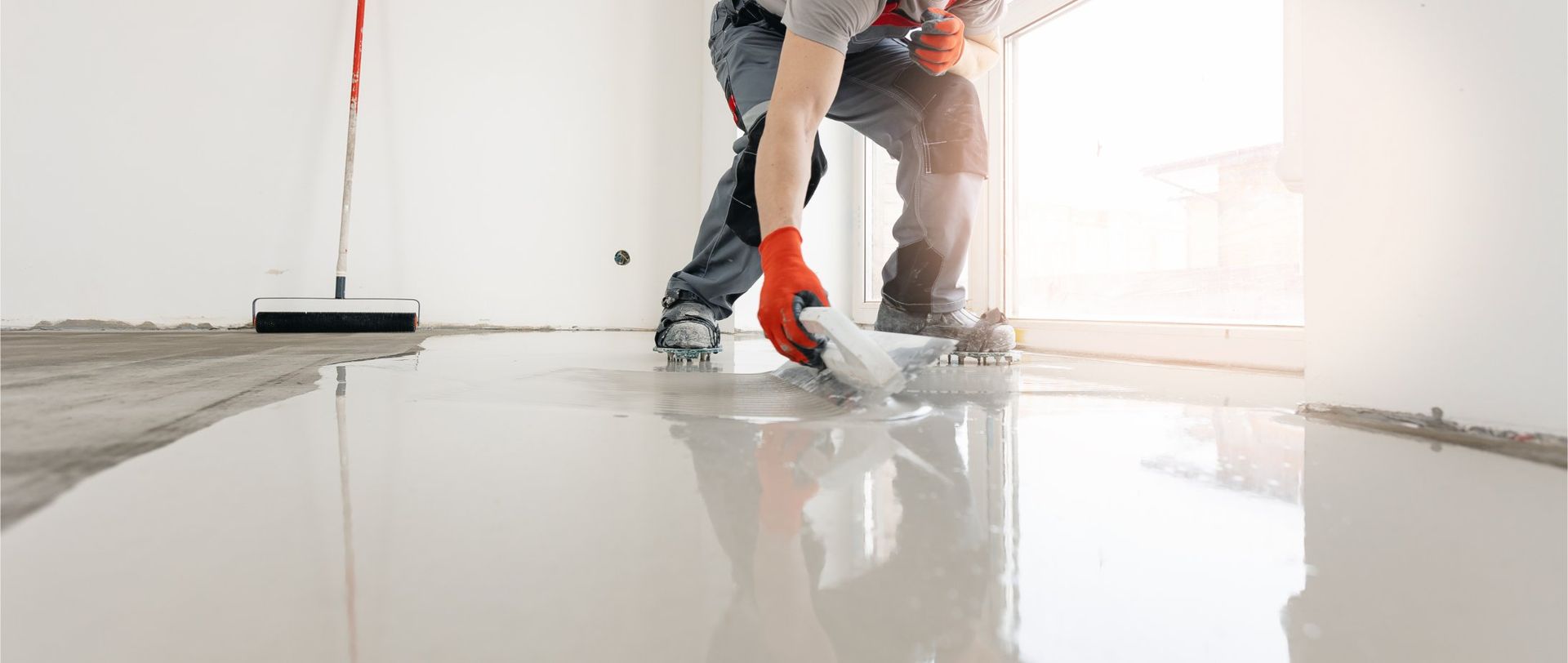 Person smoothing a light gray coating on a floor with a trowel, wearing red gloves, in a white room.