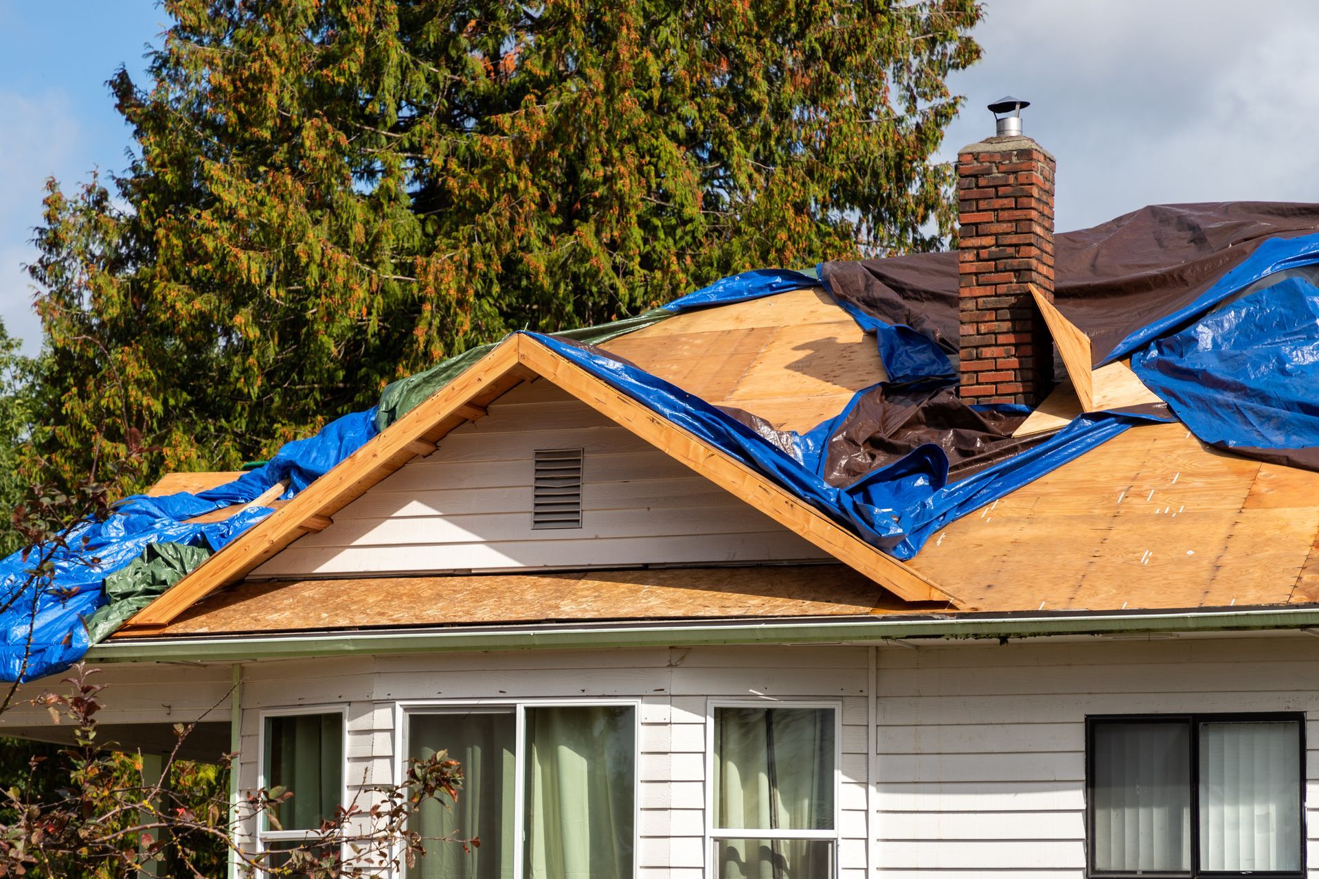 House roof under repair with blue tarp, exposed plywood, and brick chimney amid trees