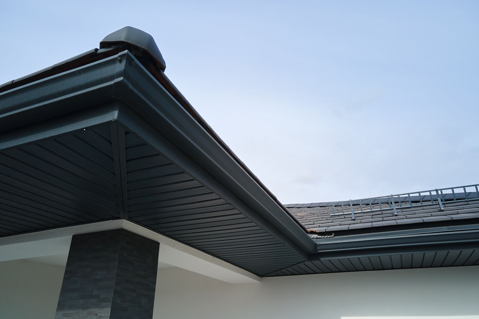 Close-up of a house roof eave with dark trim, gray siding, and a cloudy sky background