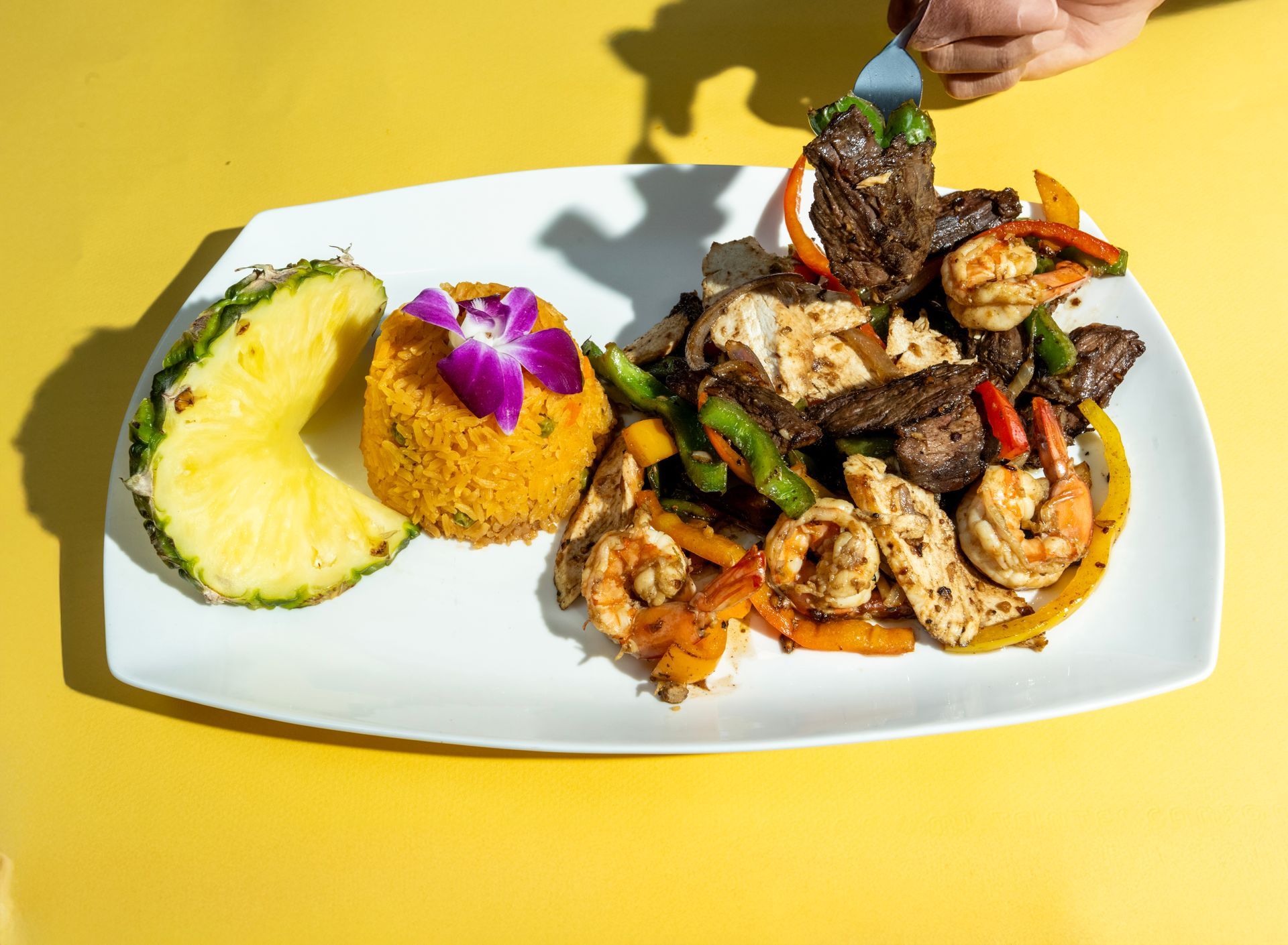 Plate of food with shrimp, meat, and rice, pineapple slice, yellow background. Hand picking up meat.