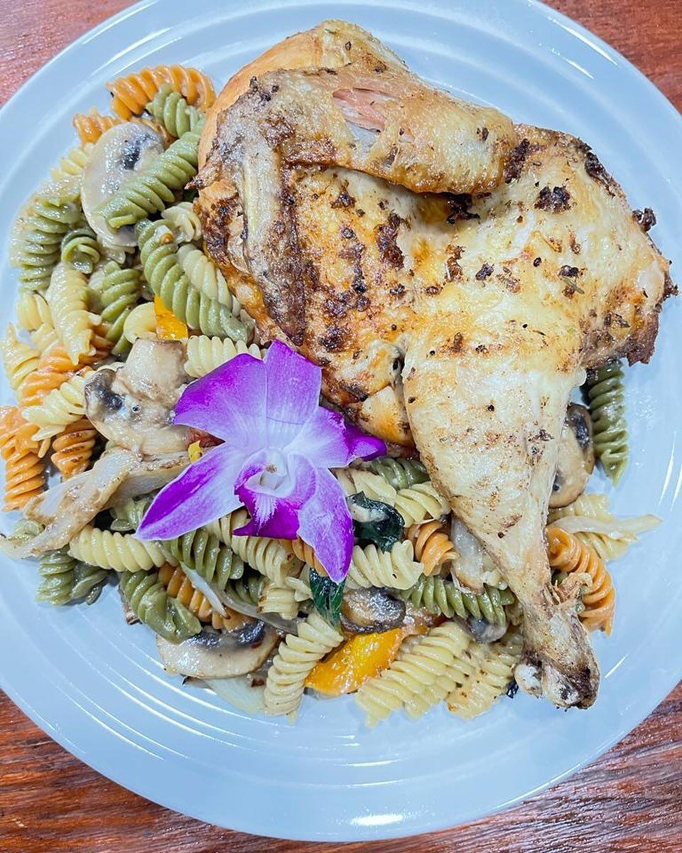 Roasted chicken quarter atop tri-color rotini pasta with mushrooms and a purple flower garnish.