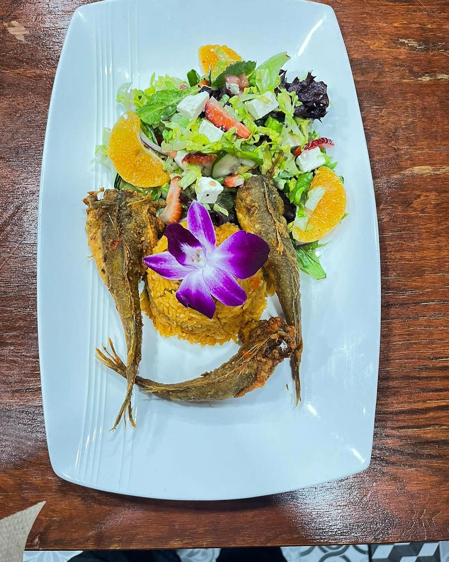 Fried fish with salad and couscous on a white plate, garnished with orange slices and a flower.