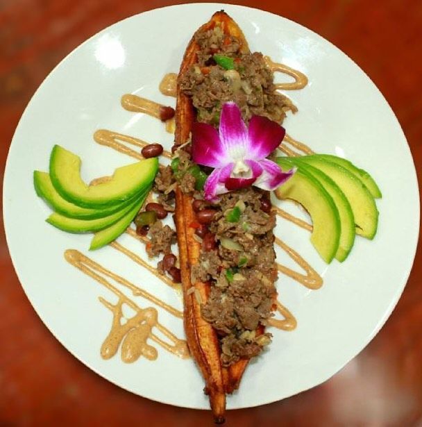 Plantain topped with meat, beans, avocado, and sauce, garnished with a flower, on a white plate.
