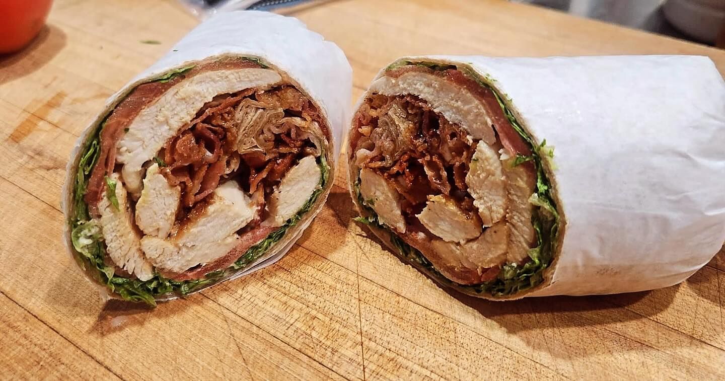 Wrap sandwich cut in half, showcasing chicken, bacon, and lettuce, wrapped in white paper on a wooden surface.